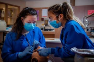 Two dental students in scrubs practice on a dummy in a classroom setting.